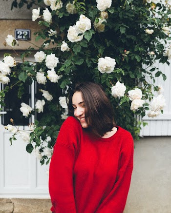 Photo of Smiling Woman in Red Long-sleeved Sweater Standing In front of White Rose Flowers