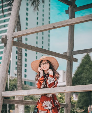 Photo of Smiling Woman in Sun Hat and Floral Dress Leaning on Wooden Structure