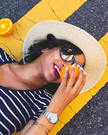 Photo of Smiling Woman In Sun Hat and Sunglasses Holding Orange While Lying Down on Concrete Road