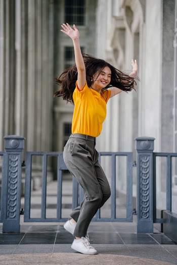 Photo of Smiling Woman in Yellow T-Shirt and Grey Pants With Her Hands Raised