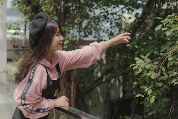 Photo of Smiling Woman Leaning Against Metal and Glass Railing Reaching Out Her Hand