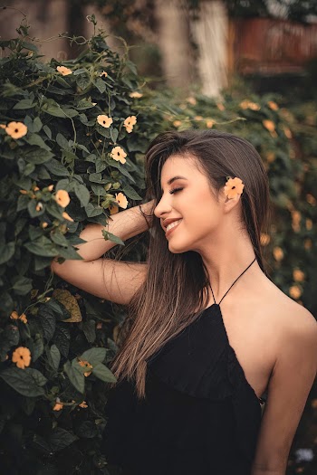 Photo of Smiling Woman with Flower on Her Hair Posing Next to Green-leafed Hedge with Her Eyes Closed