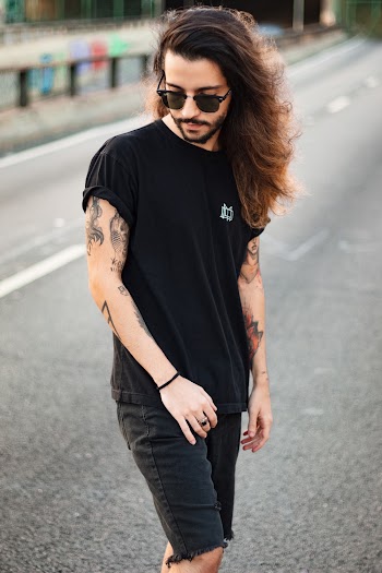 Photo of Tattooed Man in Black T-shirt, Sunglasses, and Black Denim Shorts Posing in Middle of Road While Looking Down