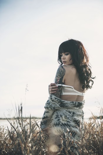 Photo of Tattooed Woman in Grey Sweater and White Bra Posing by Grass Next to Body of Water