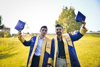 Photo of Two Men in Academic Gowns Holding Up Their Graduation Caps While Standing in Grass Field