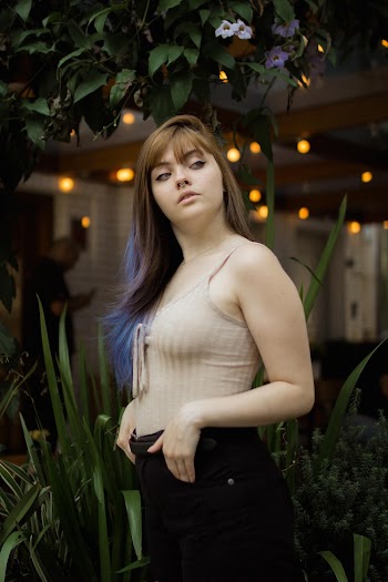 Photo of Woman in Beige Spaghetti Strap Top and Black Bottoms Standing in Front of Green Linear Leaf Plant Looking Away
