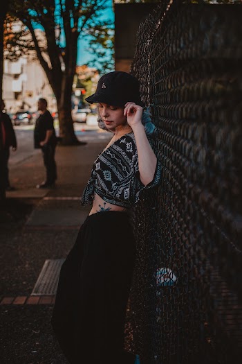 Photo of Woman in Black Crop Top, Hat, and Bottoms Leaning on Chain-link Fence by Sidewalk Posing While Looking Down