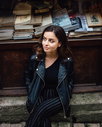 Photo of Woman in Black Leather Jacket Sitting on Stone Curb In Front of a Wooden Table with Books Posing while Looking Away