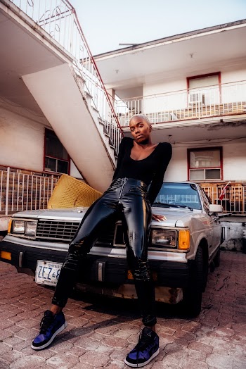 Photo of Woman in Black Leather Pants and Black Top Posing While Leaning on Hood of Parked Car