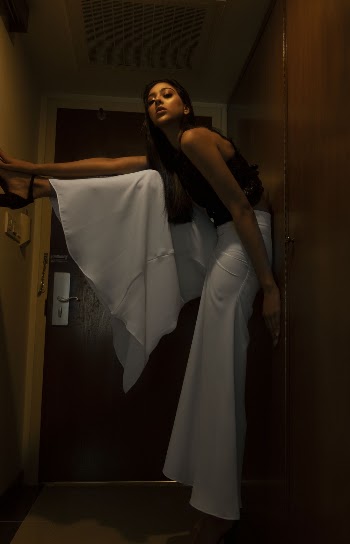 Photo of Woman in Black Top and White Palazzo Pants Standing by Doorway Posing While Leaning on Wall and Her Leg Against Wall
