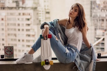 Photo of Woman in Denim Outfit Holding Penny Board While Sitting on Window Sill