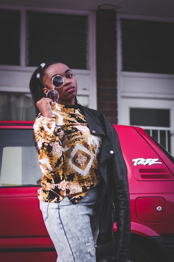 Photo of Woman in Floral Shirt and Blue Jeans Posing With Sunglasses While Standing Beside Red Car