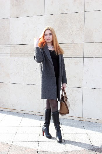 Photo of Woman in Gray Coat Posing While Holding Her Hair and Carrying a Brown Leather Bag Standing In Front of Gray Concrete Wall