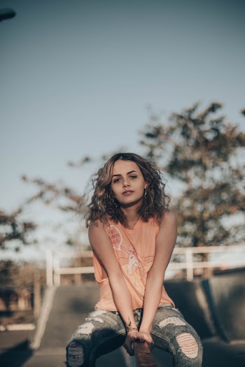 Photo of Woman in Orange Sleeveless Shirt and Blue Denim Jeans Sitting on Railing