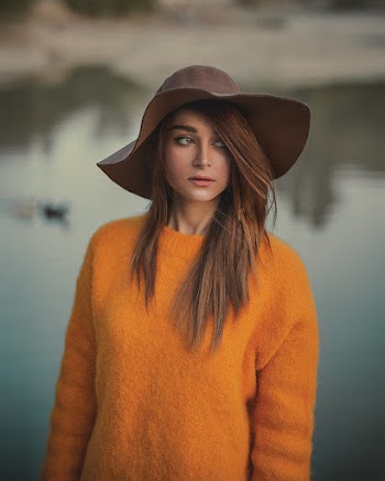 Photo of Woman in Orange Sweater and Brown Hat Posing While Looking Away with Body of Water in the Background