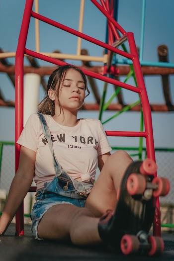 Photo of Woman in Pink T-shirt, Blue Dungarees, and Roller Skates Sitting on the Floor with her Eyes Closed With Jungle Gym in the Background