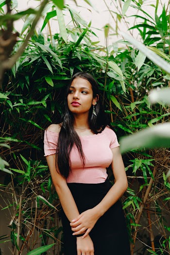 Photo of Woman in Pink Top and Black Skirt Posing In Front of Green-leafed Plants While Looking Away