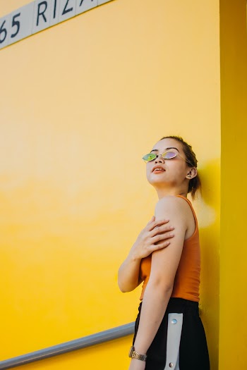 Photo of Woman in Sunglasses,Orange Top, and Black Bottoms Standing Beside Yellow Wall