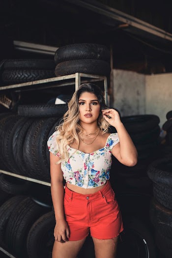 Photo of Woman in White and Blue Floral Shirt and Red Short Shorts Standing In Room Full of Tyres