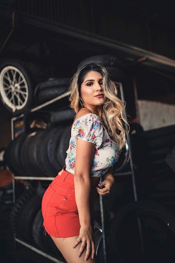 Photo of Woman in White and Blue Floral Top and Red Short Shorts Posing In Room Full of Tyres
