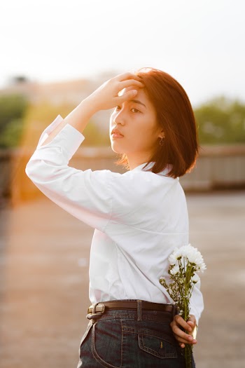 Photo of Woman in White Blouse and Blue Jeans Holding Bouquet of Flowers Behind Her Back