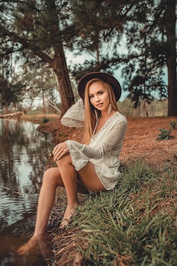 Photo of Woman in White Long-sleeved Dress and Brown Fedora Hat Sitting Near Body of Water