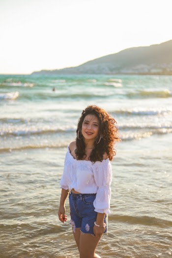 Photo of Woman in White Off-shoulder Top and Blue Denim Shorts Standing by the Beach