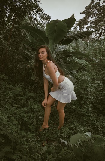Photo of Woman in White Outfit Posing Near Banana Tree Surrounded by Vegetation.