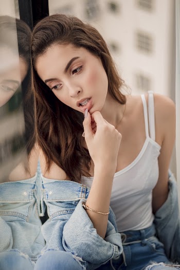 Photo of Woman in White Tank Top and Blue Distressed Denim Outfit Resting Her Head on Window While Looking Outside