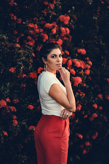 Photo of Woman in White Top and Red Pants Standing Beside Red Flowering Bush