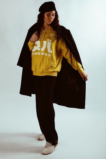 Photo of Woman in Yellow Hoodie, Black Beret Hat, and Black Coat Posing In Front of White Background