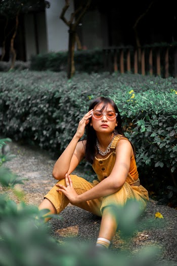Photo of Woman in Yellow Outfit and Sunglasses Sitting on the Ground Next Trimmed Bush