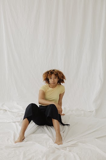 Photo of Woman in Yellow T-shirt and Black Pants Posing while Sitting on White Sheet