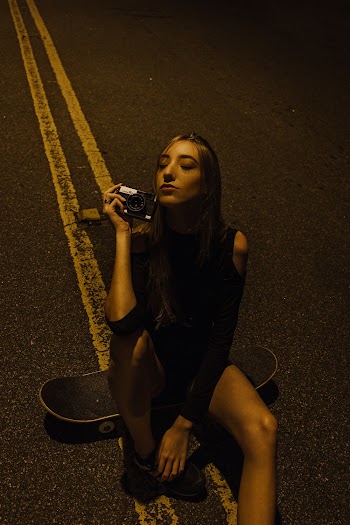 Photo of Woman Sitting on Skateboard in the Middle of the Road While Holding a Camera With Her Eyes Closed 