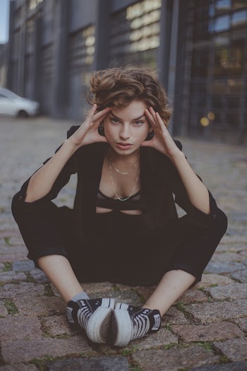 Photo of Woman Sitting on Stone Pavement While Holding Her Temple With Building in the Background