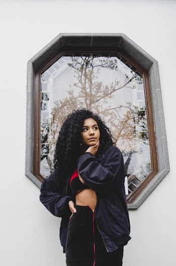Photo of Woman Wearing Black Jacket With Her Hand On Her Chin Standing In Front of Window