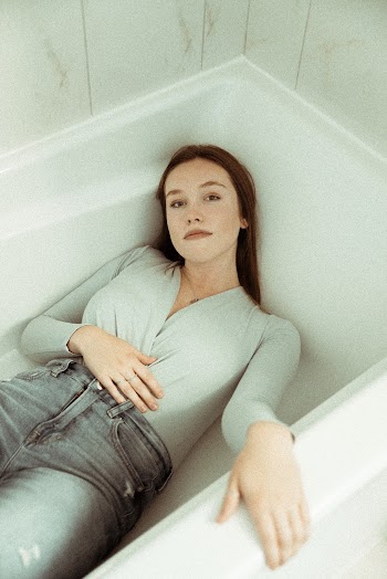 Photo of Woman Wearing Gray Long-sleeved Shirt Gray Faded Jeans Laying on White Ceramic Bathtub