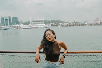 Photo of Woman With Her Eyes Closed Wearing Brown Top and Blue Denim Jeans Leaning Backwards on Wooden Railing with Harbor in the Background