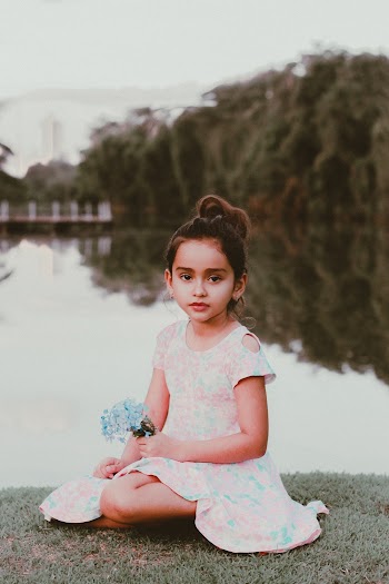 Photo of Young Girl in Make-up Wearing White Floral Dress Sitting on Grass Near Body of Water While Holding Flowers