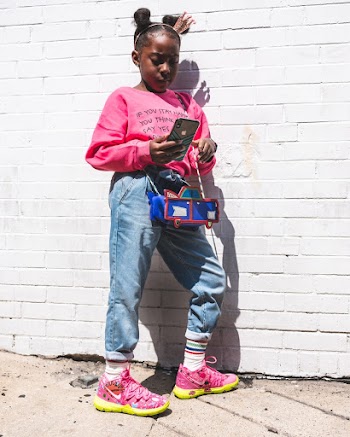 Photo of Young Girl in Pink Sweatshirt and Blue Jeans Standing and Leaning on Wall While Using a Smartphone