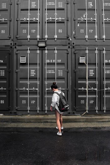 Photography of Woman in Gray Long Sleeve Shirt Carrying Black Backpack Standing in Front of Black Intermodal Container