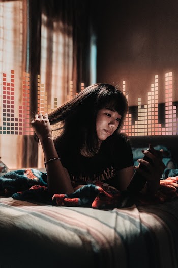 Photohraph of a woman holding her hair and looking at her phoneas she lies on the bed