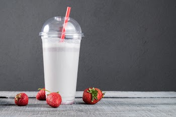 Plastic cup of milkshake with ripe strawberries on gray wooden background