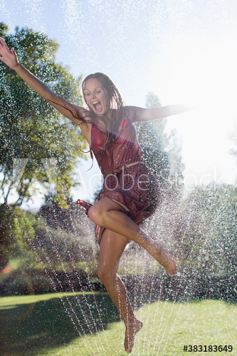 Playful young woman jumping in sprinkler in sunny backyard