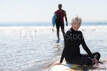 Portrait happy active senior woman sitting on surfboard in sunny surf