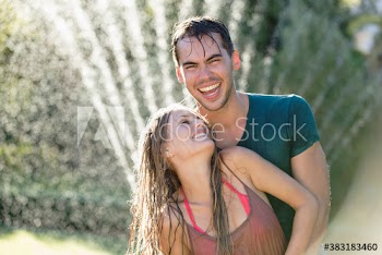 Portrait happy playful couple standing in sprinkler in sunny yard