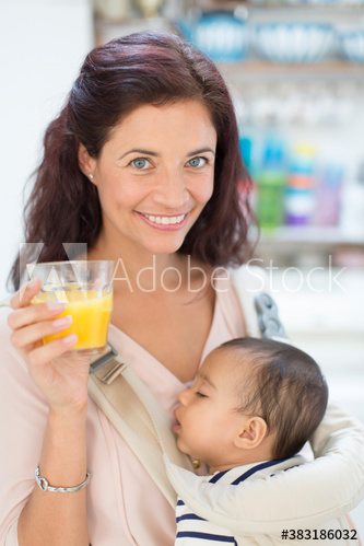 Portrait mother drinking orange juice and holding baby son in carrier