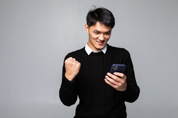 Portrait of a young asian man looks happy while reading a good news on the smartphone on white wall Free Photo