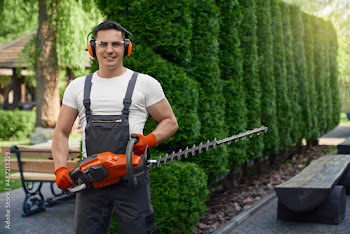 Portrait of caucasian male gardener in brown overalls, glasses and gloves holding modern hedge trimmer while posing at summer garden. Concept of people, equipment and manual work.
