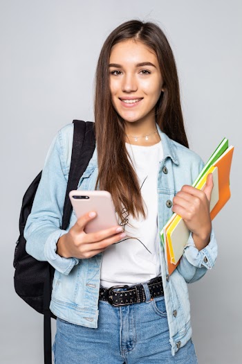 Portrait of happy young woman standing with backpack holding books and mobile phone isolated on white wall Free Photo
